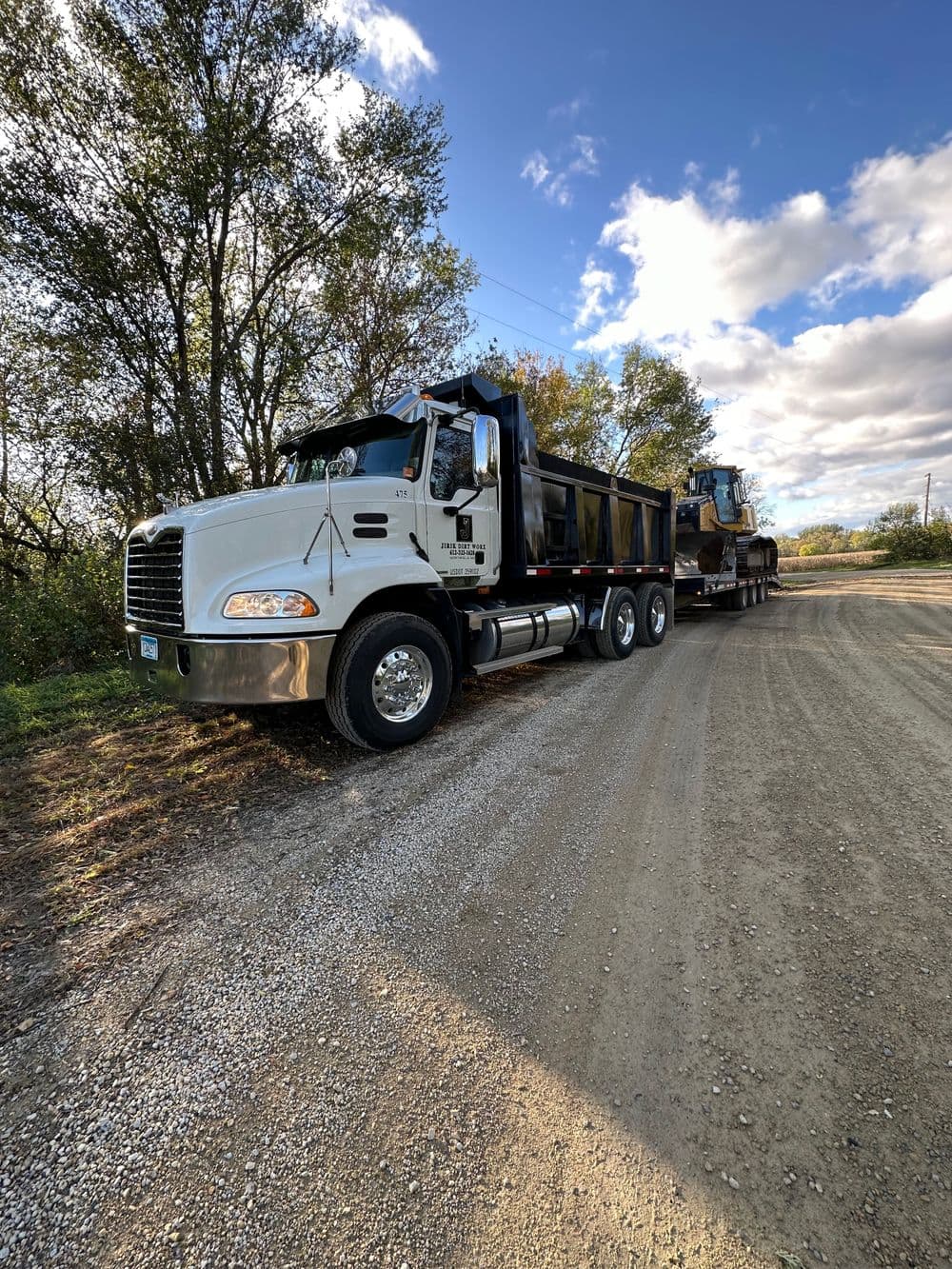 White dump truck parked on a dirt road, surrounded by trees and blue sky.