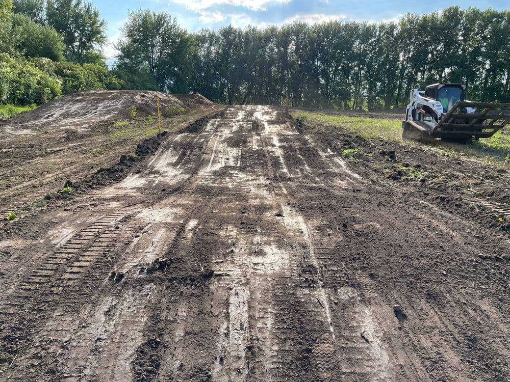 Dirt track with tire marks, truck on the side, surrounded by trees in sunny weather.