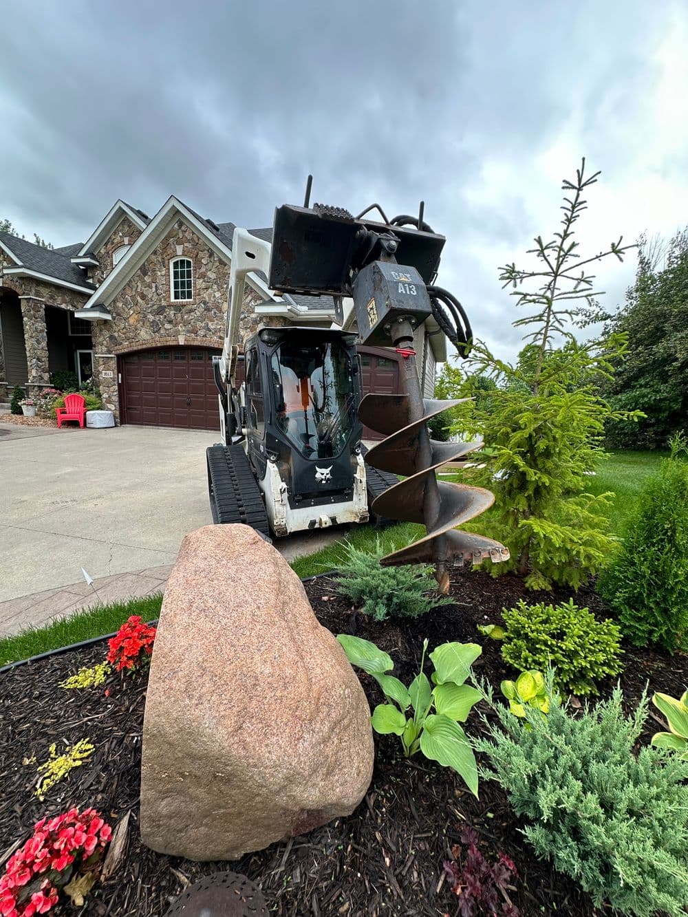 Bobcat compact excavator with auger in residential landscape, surrounded by plants and rocks.