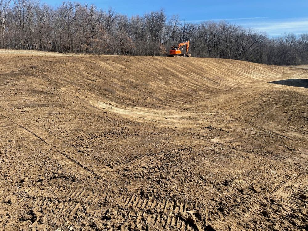 Excavator working on a cleared, dirt landscape with trees in the background.