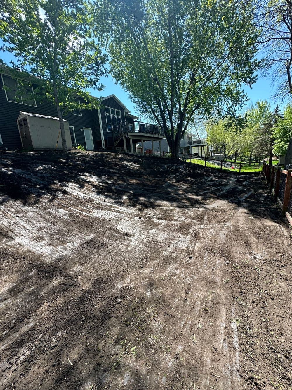 Landscape showing a sloped backyard with trees and a house in sunny weather.